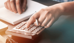 A man is using a silver calculator to do payroll for his company. He is holding a pen and his laptop is in front of him.