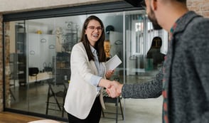 A smiling woman shakes a man's hand in an office environment. 