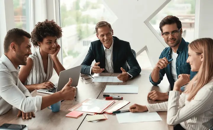 Five colleagues sat around a wooden table and smiling at each other. In front of them is a lot of paperwork, a laptop, stationery and their phones.