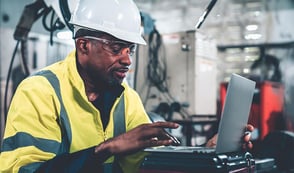 A man in a high-visibility jacket and hard hat sits at a laptop in a factory environment.