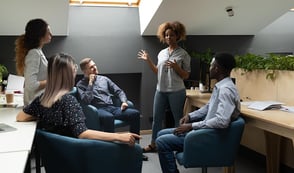 Five people are having a training meeting in a slightly dark office room. One colleague is stood up and is talking to the rest of her colleagues who are sat down and listening to her. 