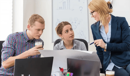 Three colleagues are sat at a desk together. They are have laptops in front of them but are all looking at a large piece of paper that has guidelines to project on.