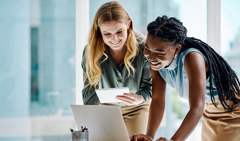 Two women look at a laptop on a table in an office environment. They are smiling. 