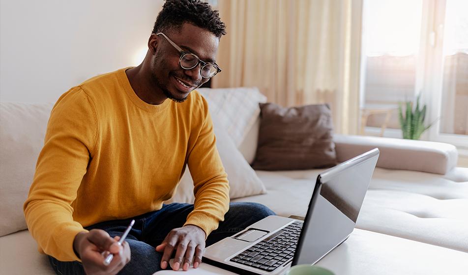 A man is working from home, sitting on his sofa. His laptop rests on the coffee table in front of him. He has a pen in his hand and is smiling, as if he's talking to someone on a remote meeting.