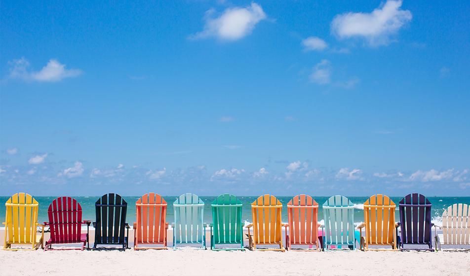 A row of multi-coloured deck chairs are lined up on a beach, facing out to sea. The photo is taken from behind them, looking out to the blue sea and the beautiful blue sky, with a few white clouds. 