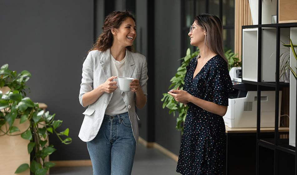 Two women stand in an office, holding coffee and smiling