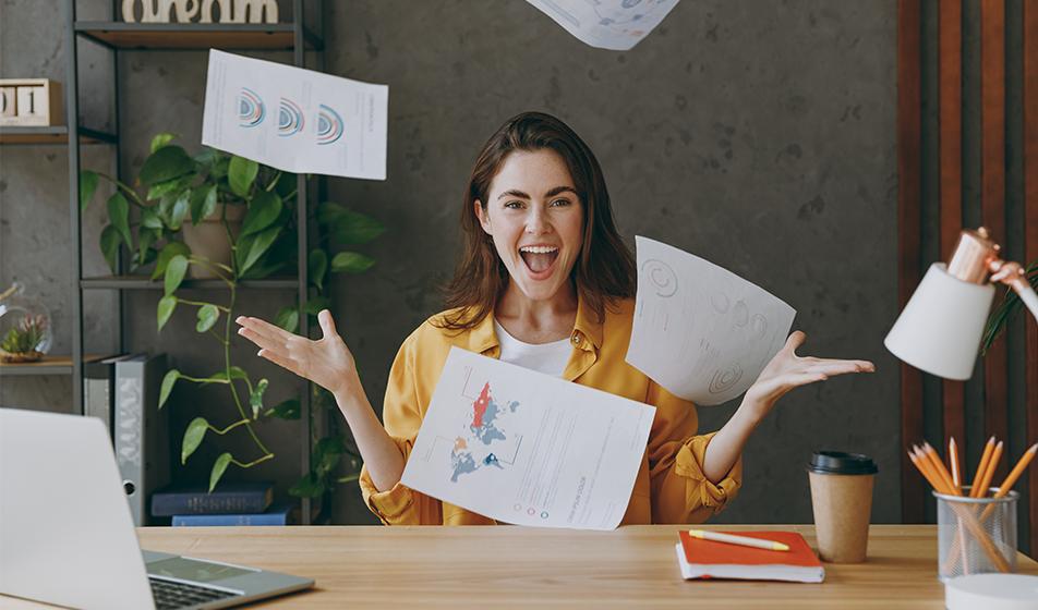 A woman sits at a desk with her laptop on. She is smiling, and has thrown 4 pieces of paperwork in the air