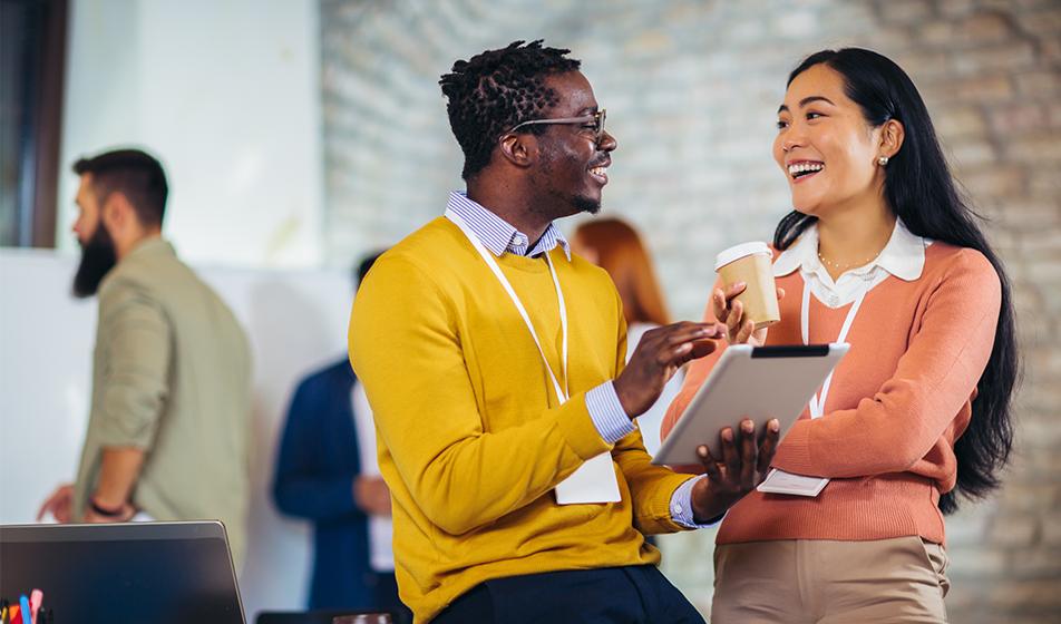 A man and a woman are chatting in an office. The woman holds a coffee, the man holds a tablet and they're smiling. 