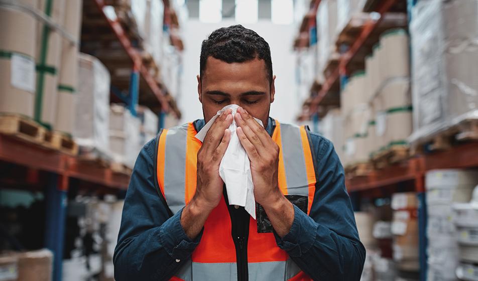 A man in a hi-vis jacket is in a warehouse. He's sneezing into a tissue. 