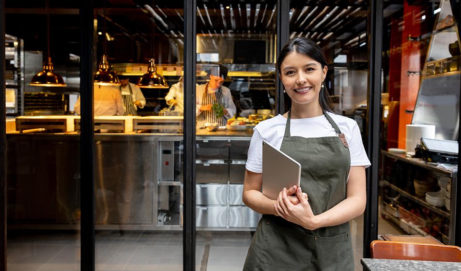 A young woman in an apron, working in hospitality, stands outside a restaurant. She's smiling & holding a clipboard. 