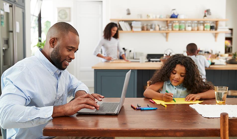 A man works at a kitchen table at his laptop, next to his daughter who's drawing with crayons. A woman and a little boy are in the background.