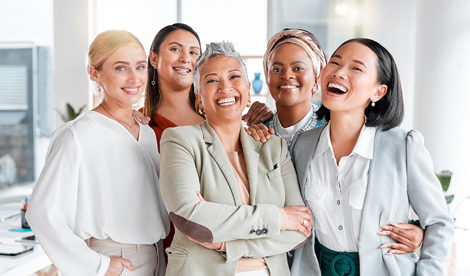 A diverse group of women of different ages stand together in a close-knit group, smiling widely.