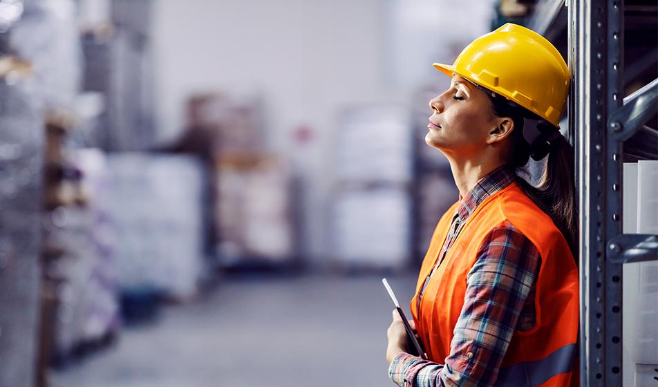 A woman in a yellow hard hat and hi-vis jacket leans against a fixture in a warehouse and closes her eyes, trying to find peace in a stressful day