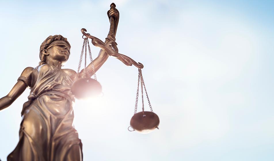 A statue representing justice is shown holding her scales of justice high against a blue sky. 