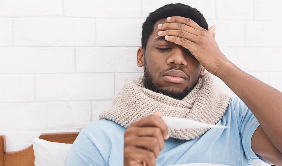 A man is feeling unwell, and has layered up with a scarf. One hand is on his forehead, as if checking temperature, and his eyes are closed. In the other hand, he's holding up a thermometer. 