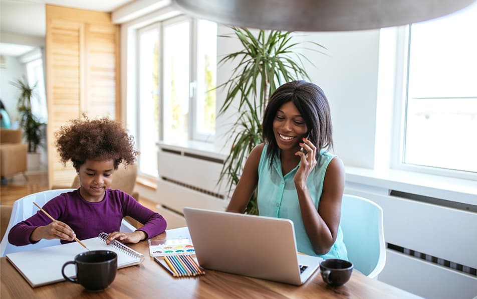 A woman and a child sat at the table together. The woman is wearing a blue top and is on the phone with her laptop in front of her. The child is wearing a purple jumper and is painting on a large piece of paper.