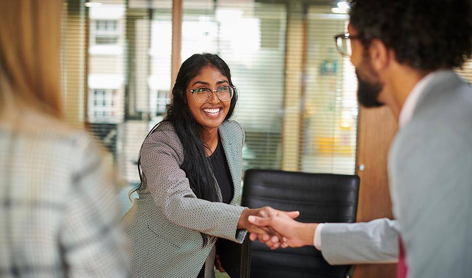A woman shakes hands across the table with one of her interviewers, smiling. The two interviewers are shown from behind.