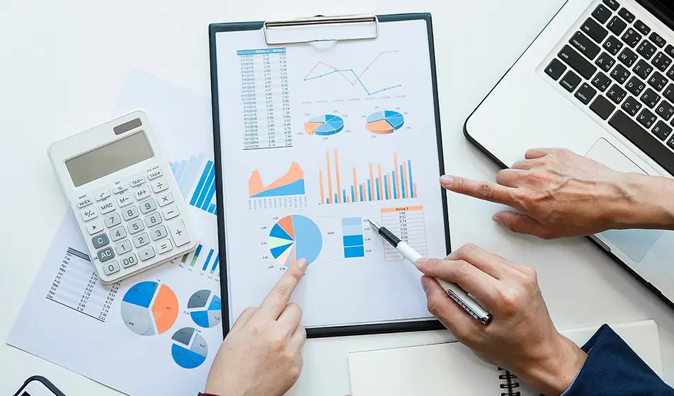 A clipboard with a piece of paper on is in the centre of the image. On the piece of paper are different types of graphs. There are three hands pointing at a graph each. The clipboard is surrounded by a laptop, a calculator and a notebook.