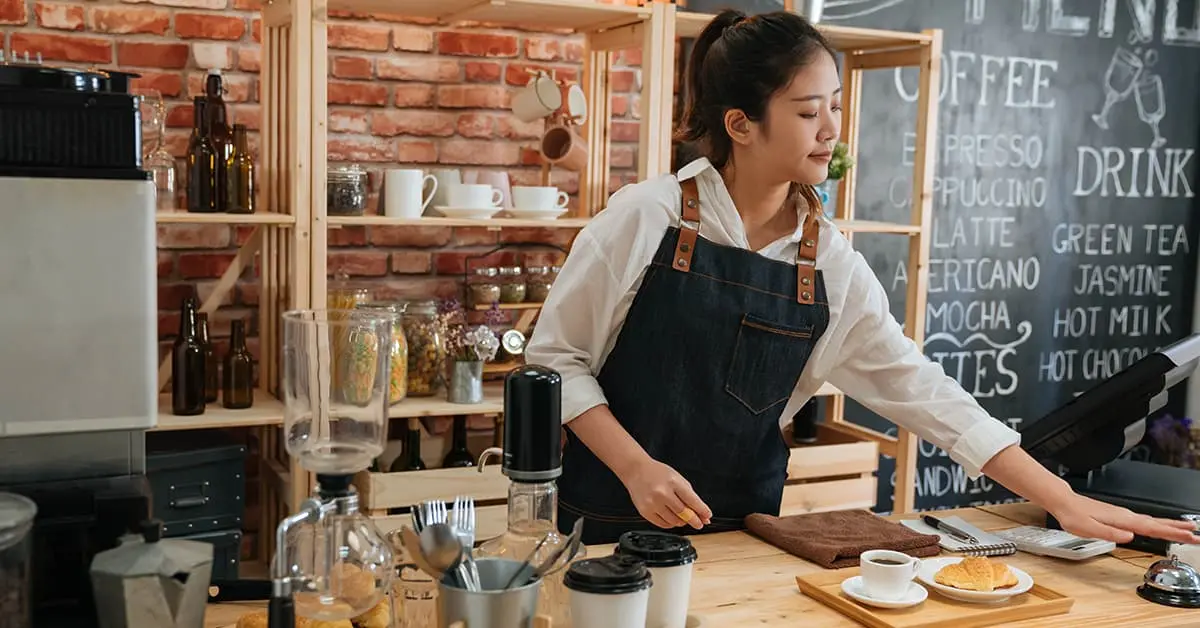 A barista working in a coffee shop is ringing the bell for someone to come and collect their order from the counter. The person has ordered a croissant and a coffee which are sat on a wooden tray.