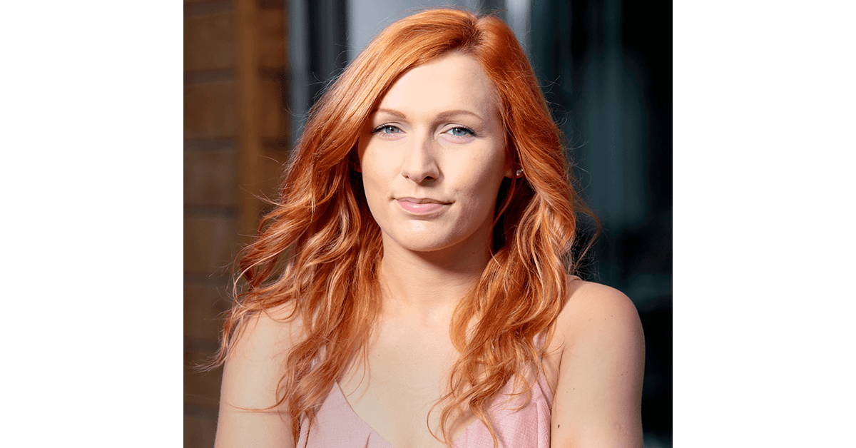 A portrait photo of a woman with bright orange hair and a pink top. She has blue eyes and is smiling at the camera.