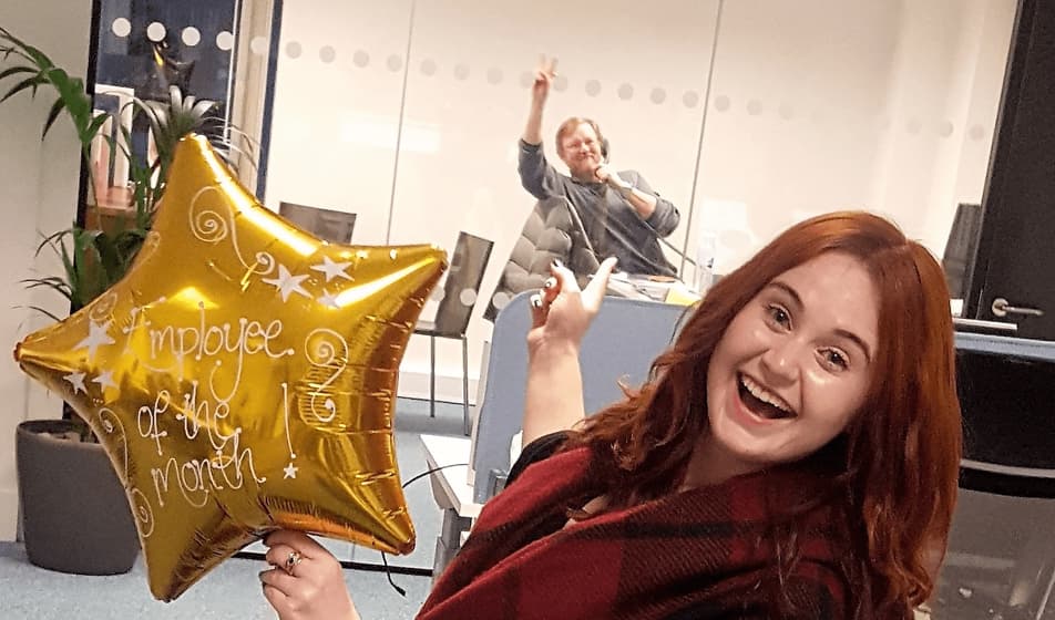 A woman is smiling and holding a star shaped golden "employee of the month" balloon. She is pointing behind her at her colleague who is on the phone in a meeting room.