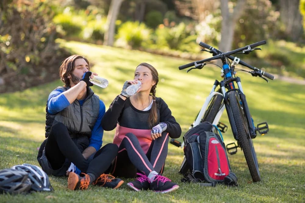 A man and a woman in cycling gear, sitting down on the grass and having a drinks break next to their bikes.