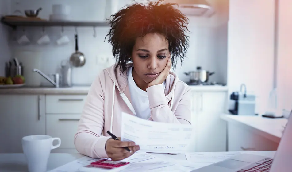 A woman is sat in her kitchen and holding a piece of paper in her hand. She is managing her finances with her laptop, a calculator  and more paperwork in front of her.