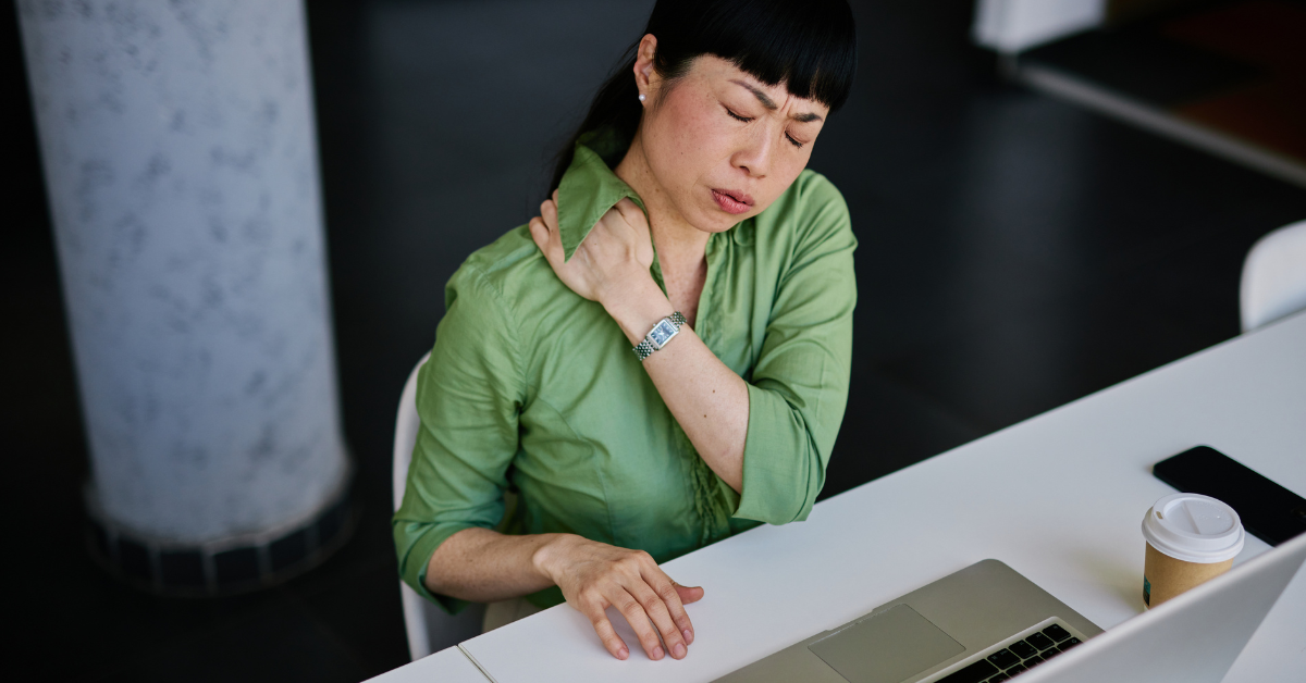 A female employee wearing glasses looks stressed while sitting at her desk, surrounded by paperwork and holding her head in her hands. Behind her, an out-of-focus colleague works at another desk. The scene reflects the pressure and confusion that can come from overlooked health and safety responsibilities in a growing business.