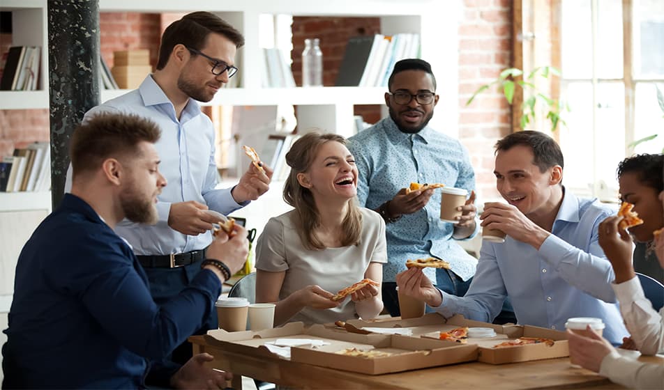 Six colleagues gathered around a table, laughing and having a pizza party.
