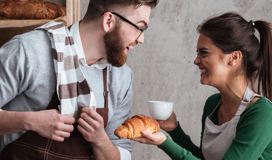 Happy woman holding a croissant and coffee laughing with her male colleague
