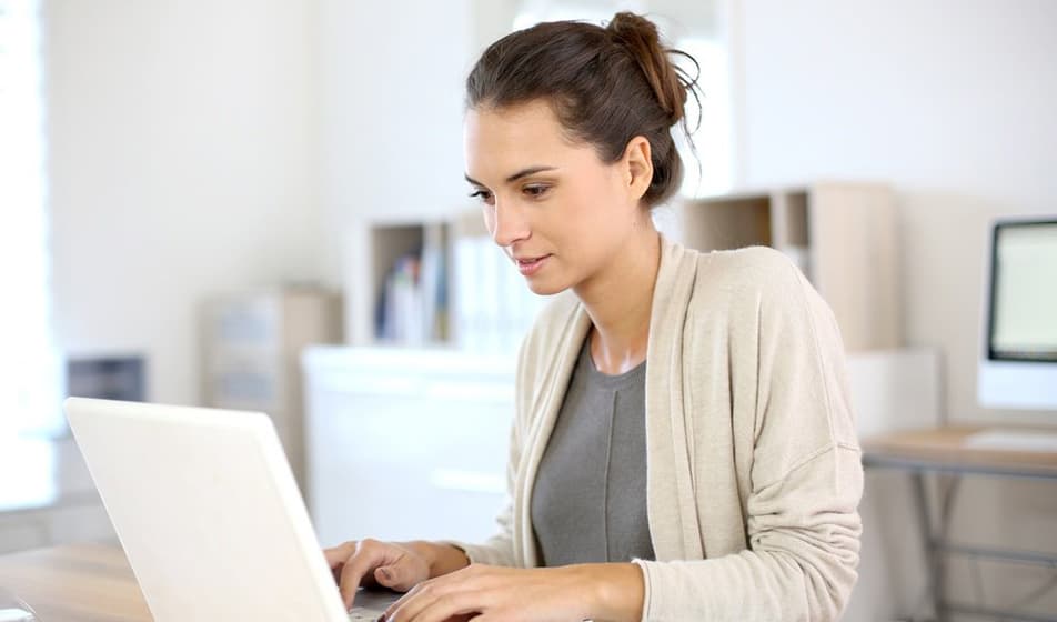 A woman with brown hair is wearing cream cardigan. She is working on her laptop and sat at a desk.