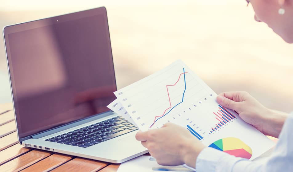 A person holding a piece of paper in front of their laptop and reviewing the bar charts on it whilst sat at her desk.