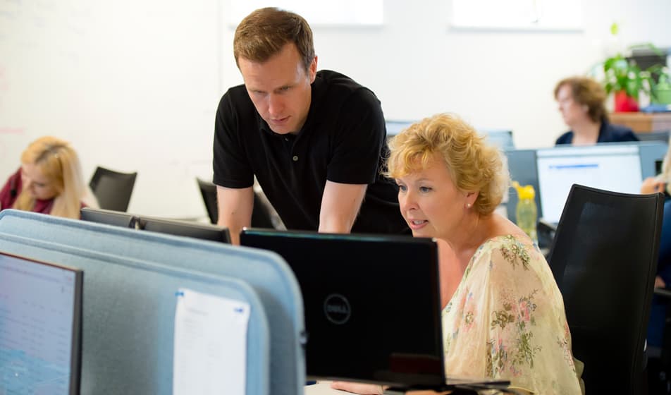 A man in a black shirt and a woman in a flowery blouse are at work and looking at a computer screen together.