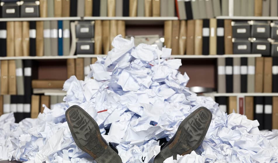 A man is covered under a pile of paperwork with only his shoes on show. He is in an office room with a wall full of files behind him.
