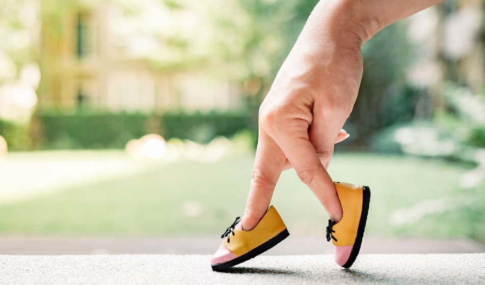 A person has two tiny yellow shoes on the ends of their fingers and is pretending that their fingers are legs, walking with these on.