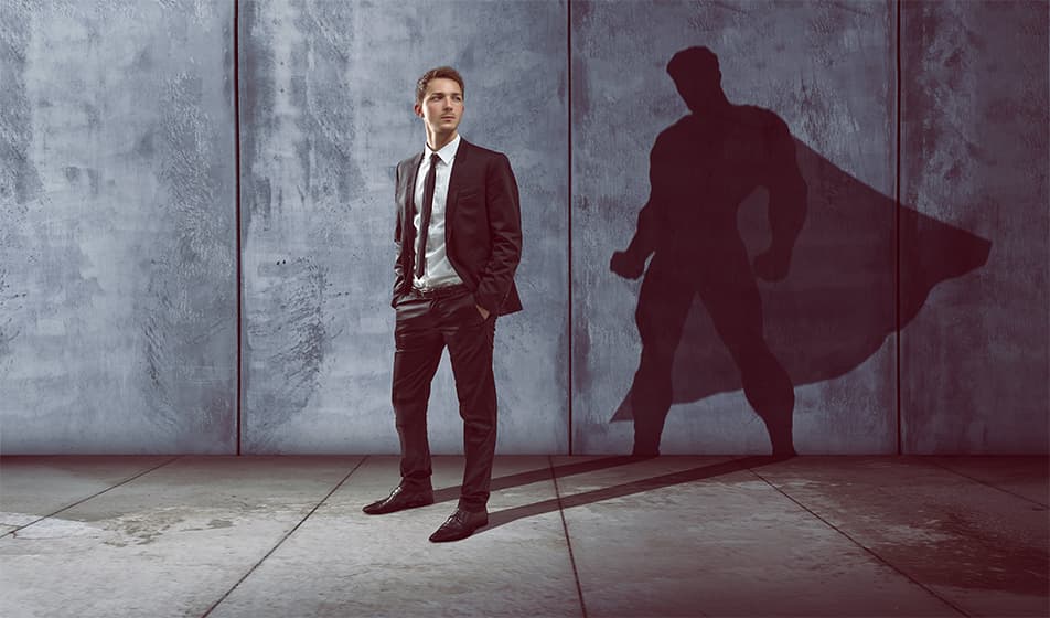 A man is in a suit and standing in a grey room. On the wall behind him is his shadow but his shadow looks like a superhero with a cape.