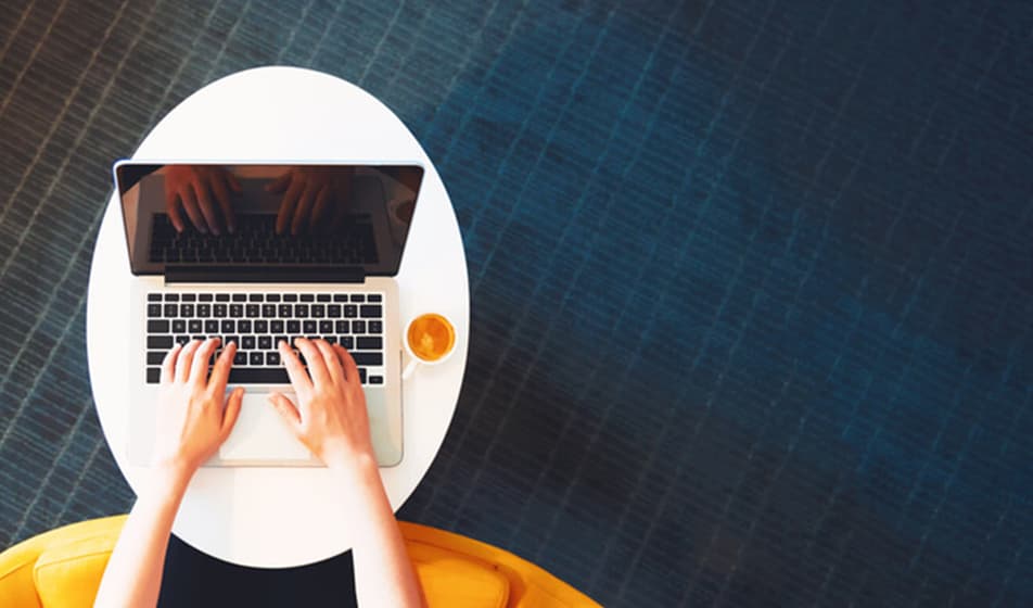 A person is sat on a yellow chair and is working on their laptop with a cup of coffee beside them.