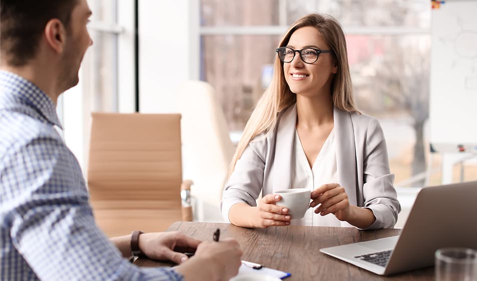 Two colleagues are sat at a table together having a meeting. The female colleague is sat next to her laptop and is smiling at her male colleague who is smiling back and writing notes.