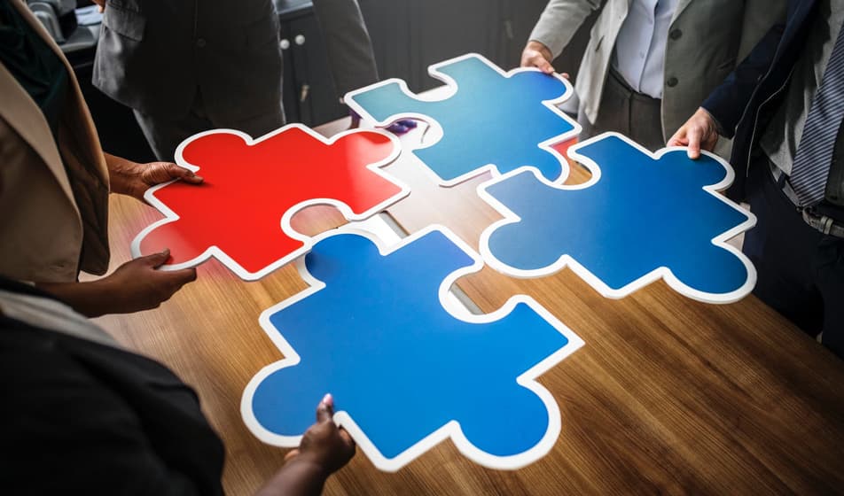 Four people are each holding one jigsaw piece. Three of the jigsaw pieces are blue and one jigsaw piece is red.