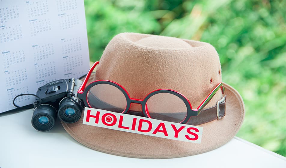 A beige hat with red and black glasses on the top of it. Also on the hat is a sign that says "holidays" which is written in red capital letters. The beige hat is sat next to some binoculars.