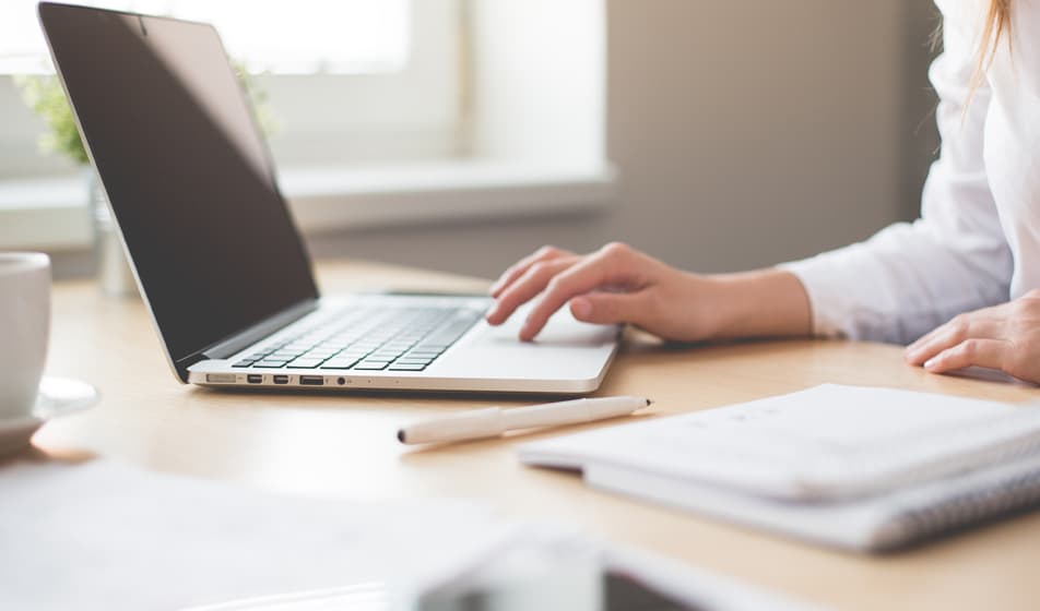 A woman in a white shirt is working at a desk on her laptop. She has her pen and notebook beside her with a white cup.