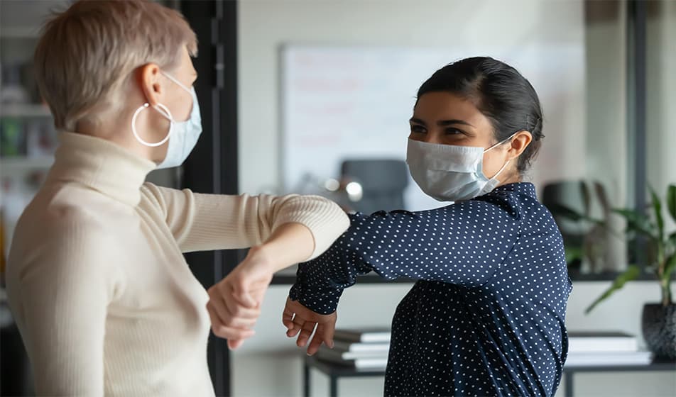 Two female colleagues are wearing masks during the pandemic. They are bumping elbows in an office.