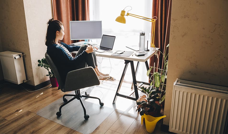 A woman is working from home and is sat with her feet up on her chair. Her chair is next to her desk which has a large window in front of it. She has one large screen next to her laptop with a yellow lamp hanging over her desk.
