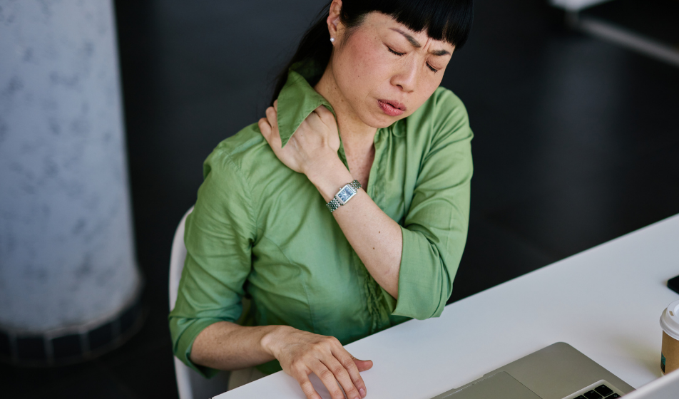 A female employee wearing glasses looks stressed while sitting at her desk, surrounded by paperwork and holding her head in her hands. Behind her, an out-of-focus colleague works at another desk. The scene reflects the pressure and confusion that can come from overlooked health and safety responsibilities in a growing business.