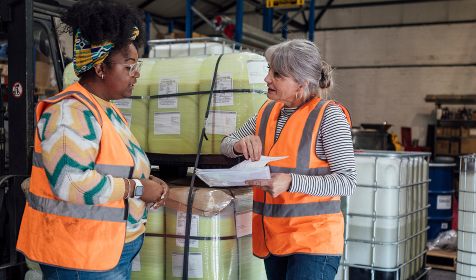 Two employees in high-visibility vests review paperwork together in a warehouse, focusing on workplace health and safety.
