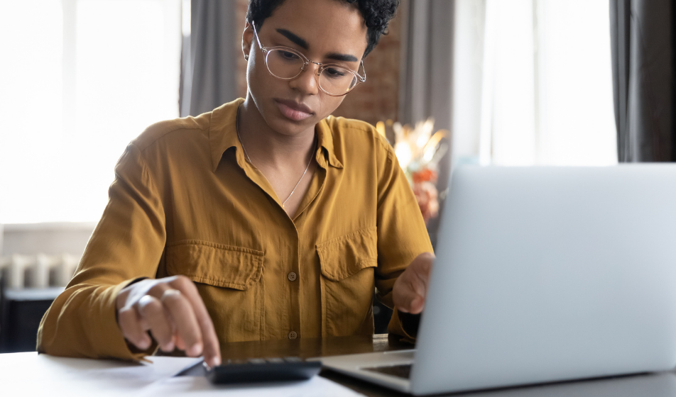 A female small business owner with short hair and round glasses, wearing a mustard yellow shirt, sits at a desk. She's pressing buttons on a calculator with one hand while referencing documents with the other, calculating employer national insurance contributions with a laptop open in front of her.