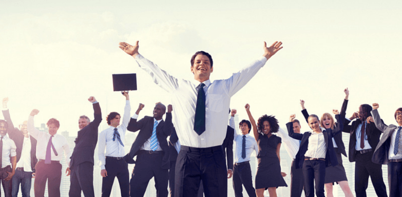 A man is standing with his hands in the air with a large line of people behind him. They are all wearing suits and also have their hands in the air celebrating.