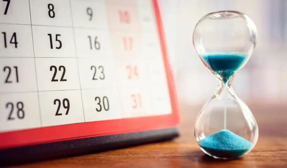 A sand timer with blue sand is stood next to a calendar. The timer is about halfway done.