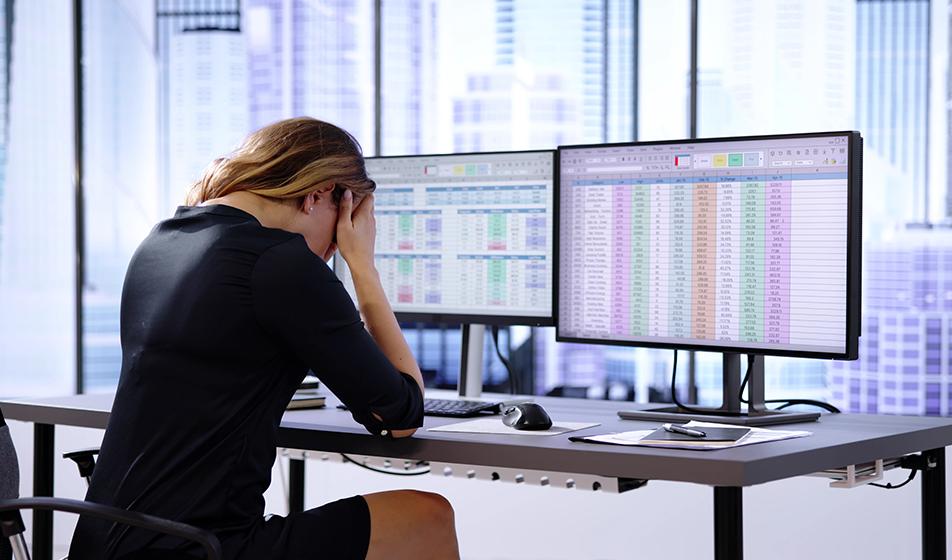 A woman sits at her desk in an office environment with her head in her hands, looking dismayed. In front of her on two computer screens are spreadsheets, showing rows and rows of data. 