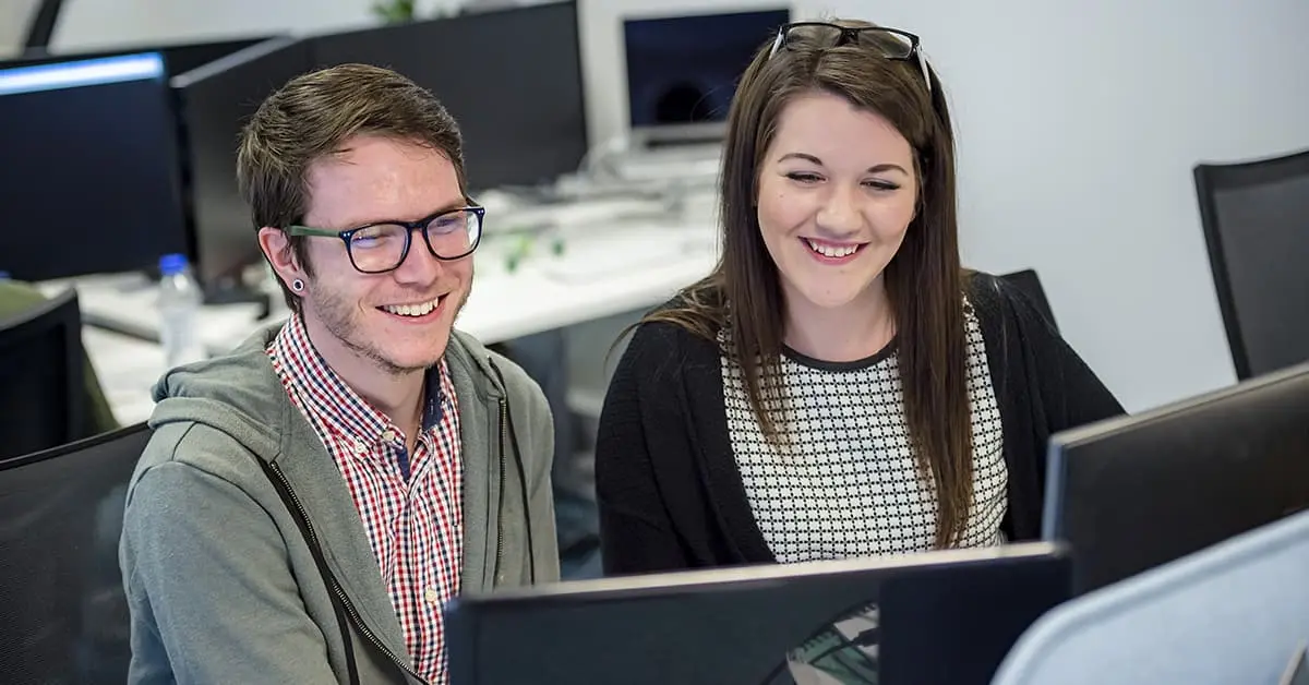 Two colleagues are sat at a desk together and looking at a computer screen. They are both smiling.